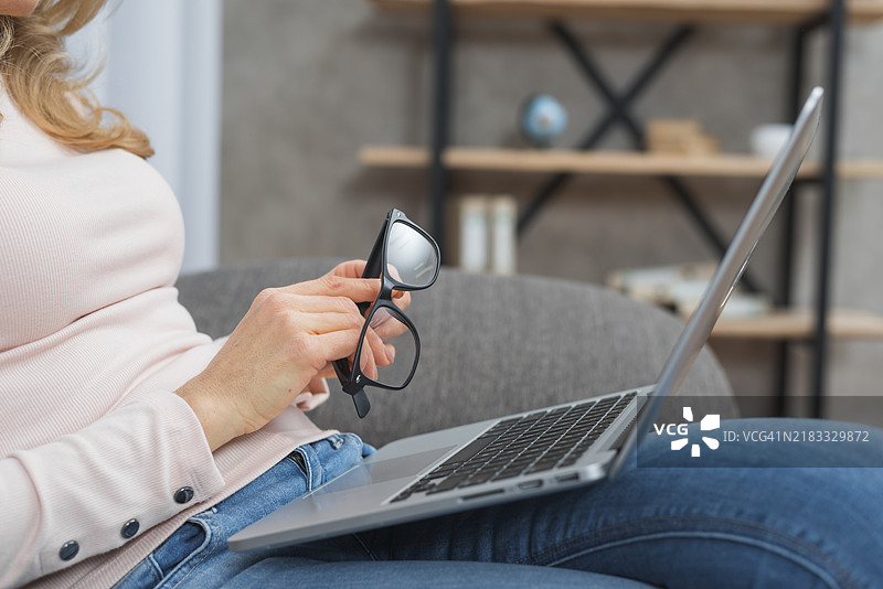 女人坐在沙发上，手里拿着眼镜，膝盖上放着打开的笔记本电脑。图片素材