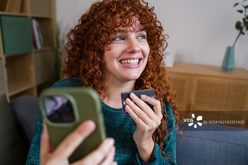 快乐的红发女性在家中使用智能手机和信用卡进行在线购物图片素材