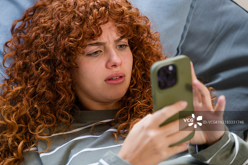 年轻女子红发，面露困惑之色，正在阅读手机信息。图片素材