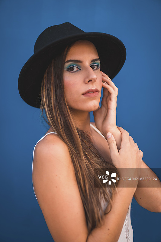 一位年轻的棕发女孩戴着黑色帽子，穿着白色T恤，背景是蓝色的。蓝色时尚妆容带有闪光效果。图片素材