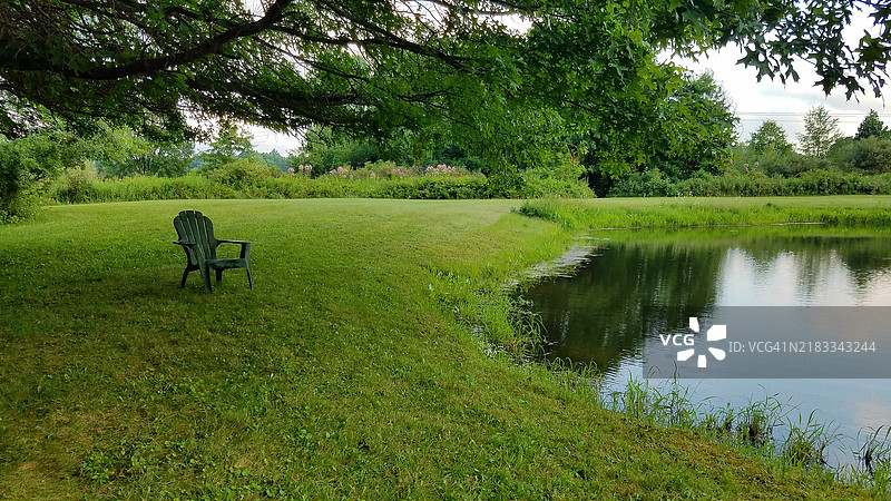 空椅子放在草地上，位于大树下，靠近池塘边缘图片素材