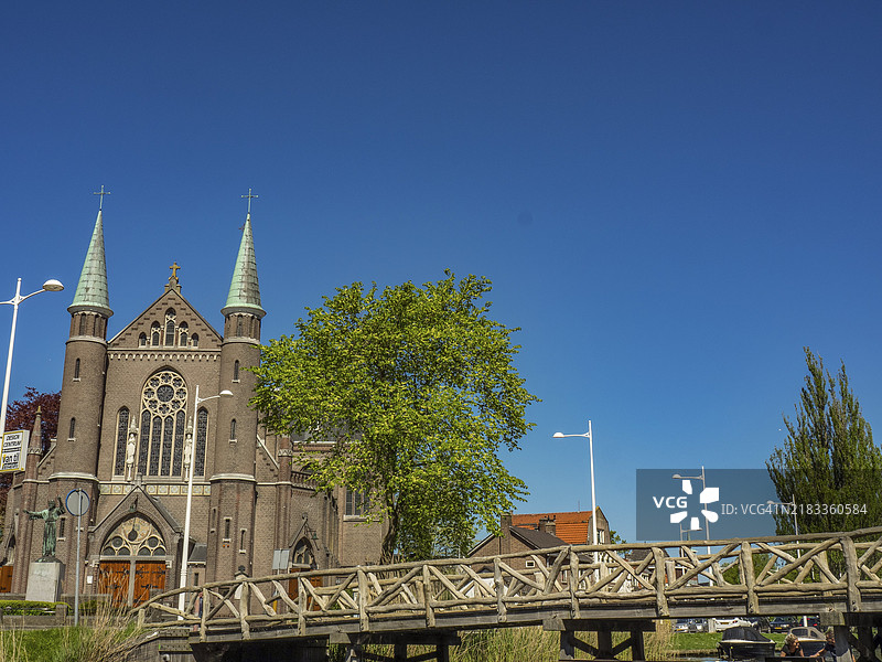 荷兰阿尔克马尔的教堂，前景有一座木桥，周围环绕着树木和湛蓝的天空。图片素材