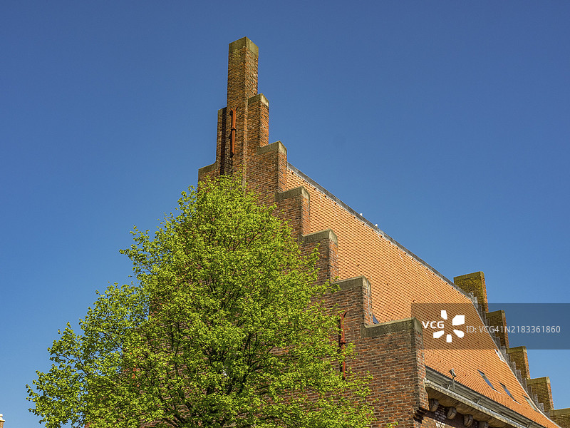 荷兰阿尔克马尔一座砖砌建筑，带有阶梯式屋顶，前景是一棵树，背景是晴朗的天空。图片素材