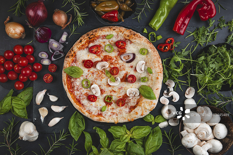 装饰有番茄和罗勒的比萨，周围环绕着新鲜的食材，如蘑菇、洋葱、芝麻菜和香草图片素材