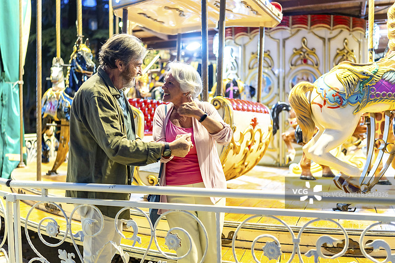 一对老年男女在古老的旋转木马上享受骑行，站在一旁聊天。图片素材