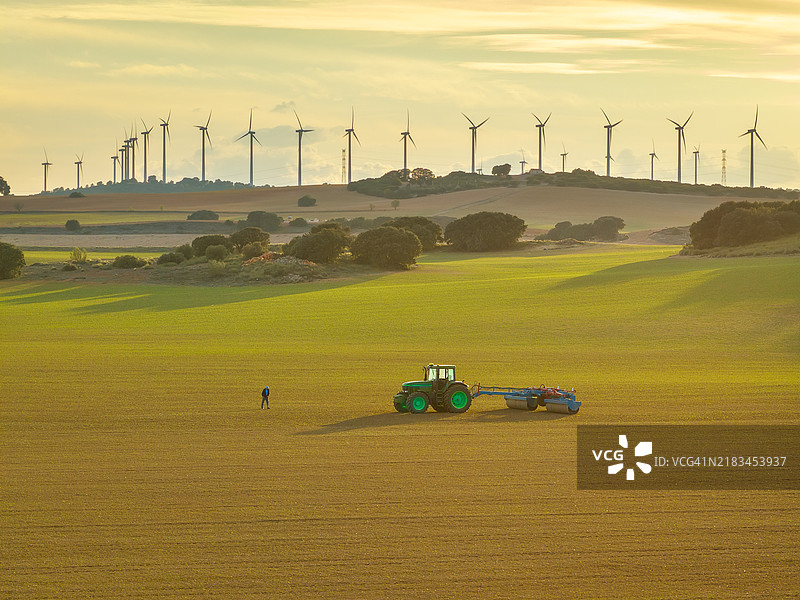 无人机航拍的风景如画的乡村景观，一位孤独的农民在日落时分与他的拖拉机一起耕作广阔的田野，远处的山脉上有一排大型风力涡轮机在旋转。西班牙图片素材