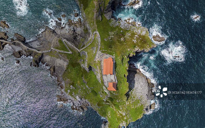 无人机拍摄，俯瞰被海浪环绕的岩石岛屿，岛上有一座红色屋顶的建筑和蜿蜒的小径，位于西班牙巴斯克地区的圣胡安·德·加斯特卢盖特。图片素材