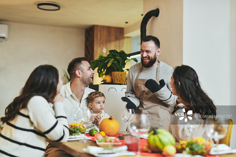 年轻家庭坐在餐桌旁，成年男性戴着厨房手套开玩笑并大笑。图片素材