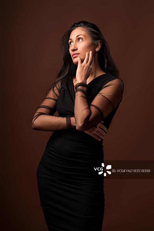 一位哥伦比亚女性优雅地穿着黑色连衣裙，在摄影棚背景前轻轻触碰自己的脸。图片素材