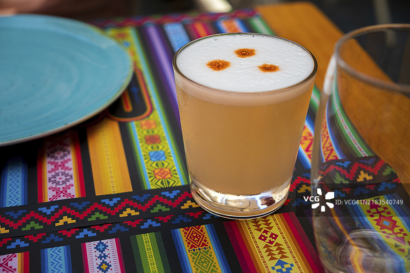 秘鲁鸡尾酒皮斯科酸酒，采用皮斯科（秘鲁葡萄白兰地）、青柠汁、糖浆和蛋白制成，盛在带有传统图案的餐垫上的玻璃杯中。选择性对焦，背景模糊，前景的玻璃杯和泡沫清晰可见。图片素材