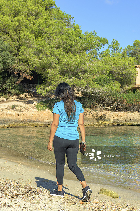 穿着运动服的拉丁裔女性在海滩上走路，边听音乐边使用智能手机图片素材