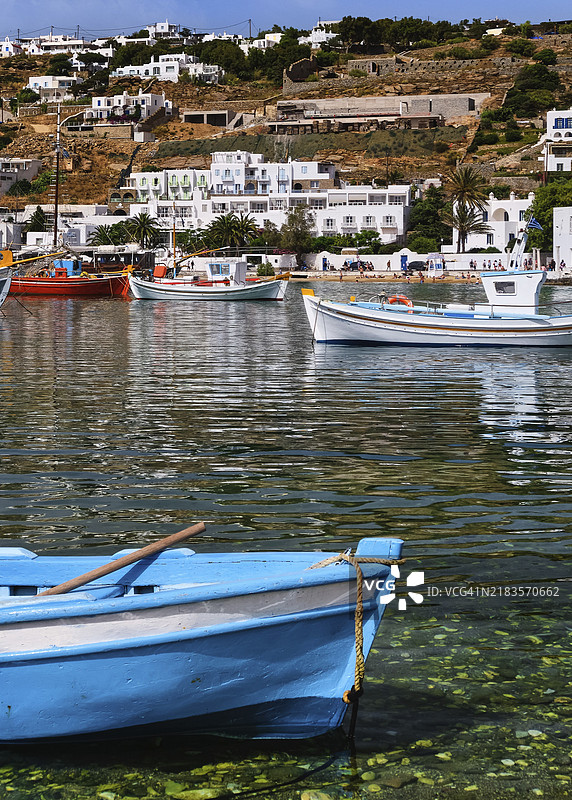 美丽的夏日阳光照耀着希腊岛屿的港口。五彩斑斓的渔船停靠在码头。海滨的山丘上是白色的房屋。地中海的生活方式,假期。前景中有一只小蓝船。米科诺斯,基克拉泽斯群岛,希腊。竖拍。图片素材