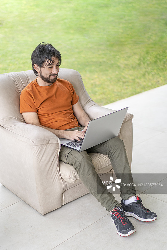 拉丁裔男子坐在沙发上，在花园里用笔记本电脑工作图片素材