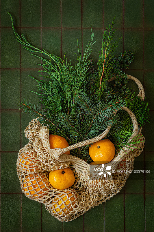 成熟的橘子装在绳网袋中，旁边有云杉枝，背景为绿色。这是圣诞节假期的概念。图片素材