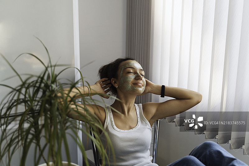 戴着面膜的女人在阳光下的窗边放松。图片素材