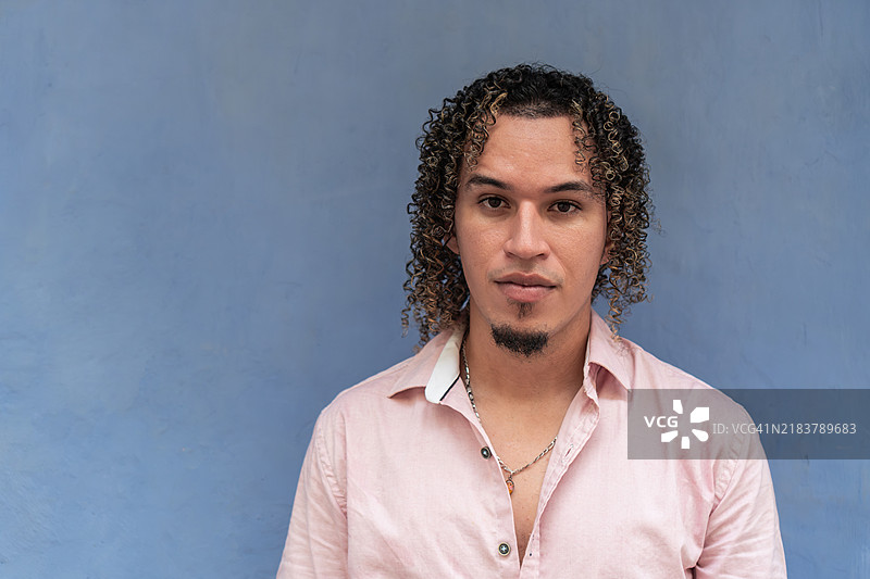 穿着粉色衬衫的卷发西班牙裔男子站在蓝色墙壁前图片素材