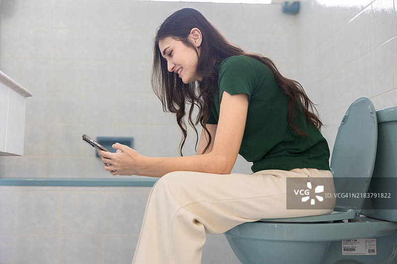 愉快的年轻女性坐在马桶上使用智能手机，营造轻松的居家环境，捕捉真实的日常生活、个人舒适以及科技在现代生活中的角色。图片素材