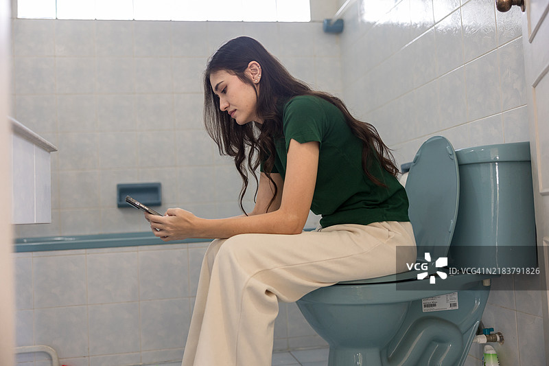 愉快的年轻女性在马桶座上使用智能手机，营造轻松的居家环境，捕捉真实的日常生活、个人舒适以及科技在现代生活中的角色。图片素材