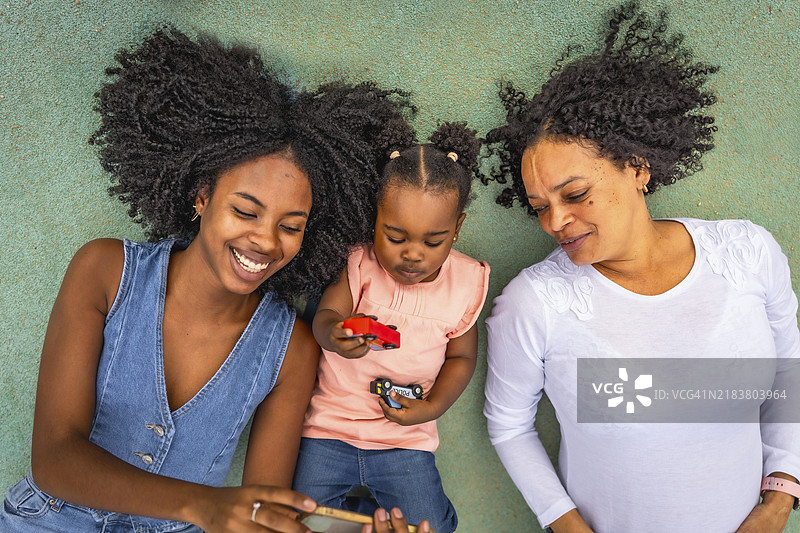 多代非洲女性在公园里躺着使用手机和玩耍。家庭概念图片素材