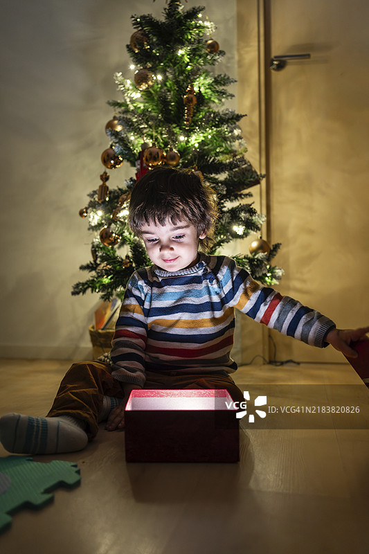 一个小男孩坐在圣诞树前的地板上，正在打开一个红色的盒子。图片素材