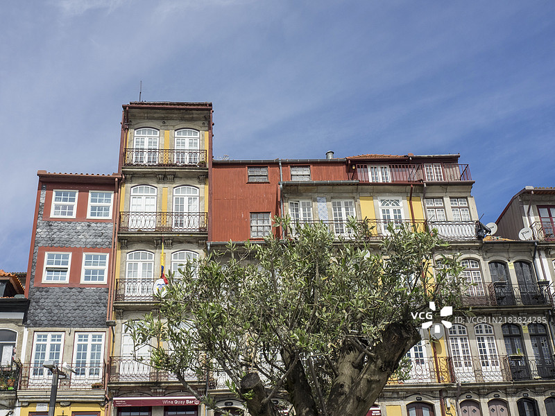 装饰华丽的老城区房屋，带有小阳台和白色窗户，蓝天之下，前景有一棵绿树，河边的彩色房屋与五彩斑斓的船只和一座大桥，波尔图，葡萄牙，欧洲图片素材