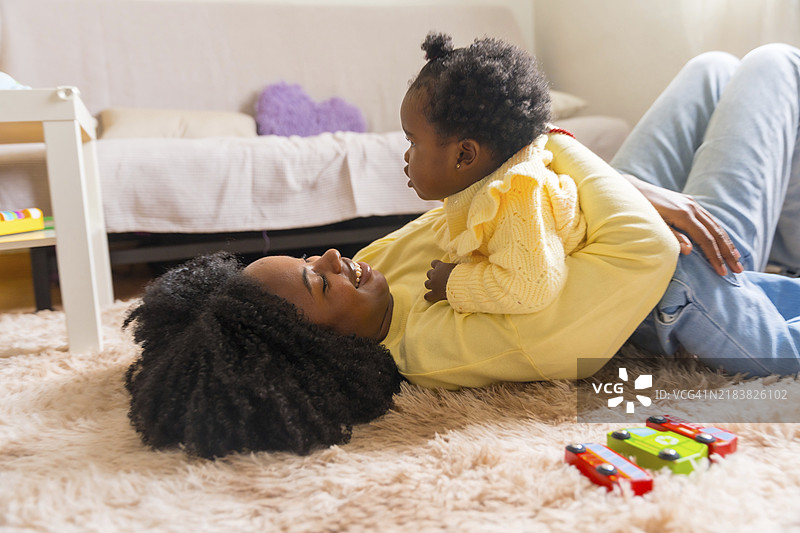 快乐而微笑的非洲女性躺在客厅的地板上，拥抱着她的女儿。图片素材
