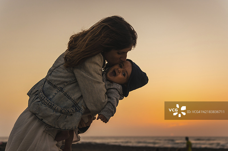 年轻母亲在西班牙加那利群岛富埃特文图拉岛的埃尔科蒂略镇附近的托斯顿灯塔日落时亲吻她的儿子。图片素材