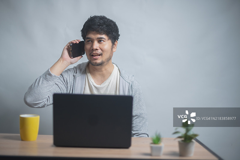 男子在笔记本电脑前打电话，桌上有一个黄色杯子和小植物，展现出轻松的工作环境或家庭办公室。图片素材