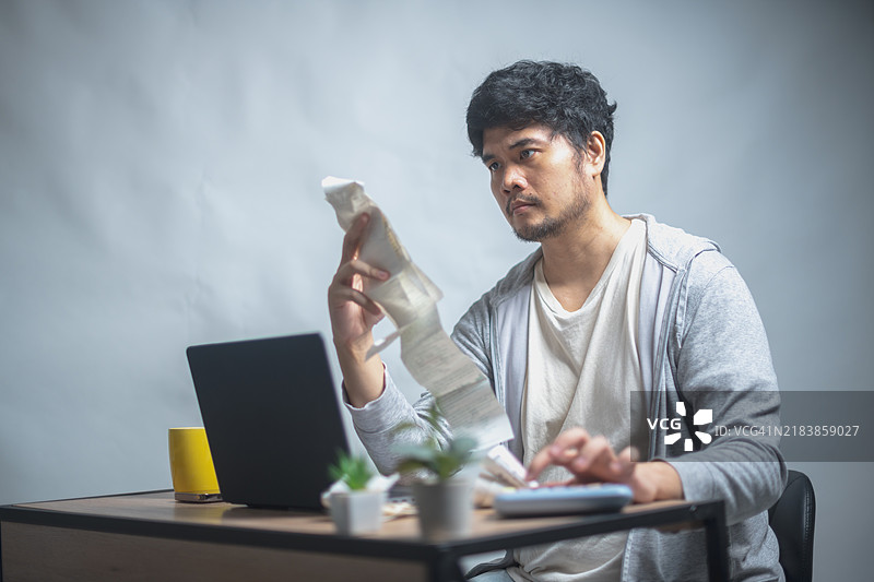 男子坐在桌子前，面露困惑地查看收据，桌上有一台笔记本电脑、一台计算器和一个黄色杯子，可能在管理财务。图片素材