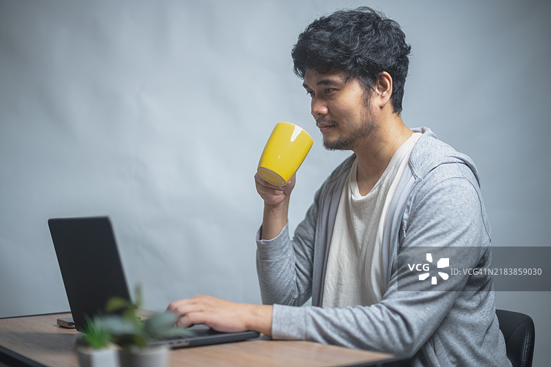 一个人坐在桌子前，使用笔记本电脑，一边打电话，一边喝着黄色的杯子，旁边有一盆植物。图片素材