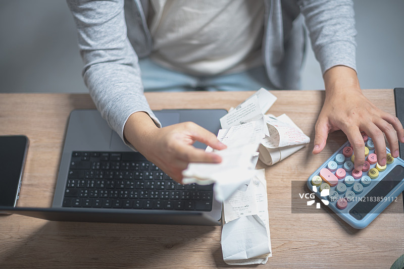 一个人正在用笔记本电脑和计算器进行预算或会计工作，周围是一堆皱巴巴的收据，散落在木质表面上。图片素材