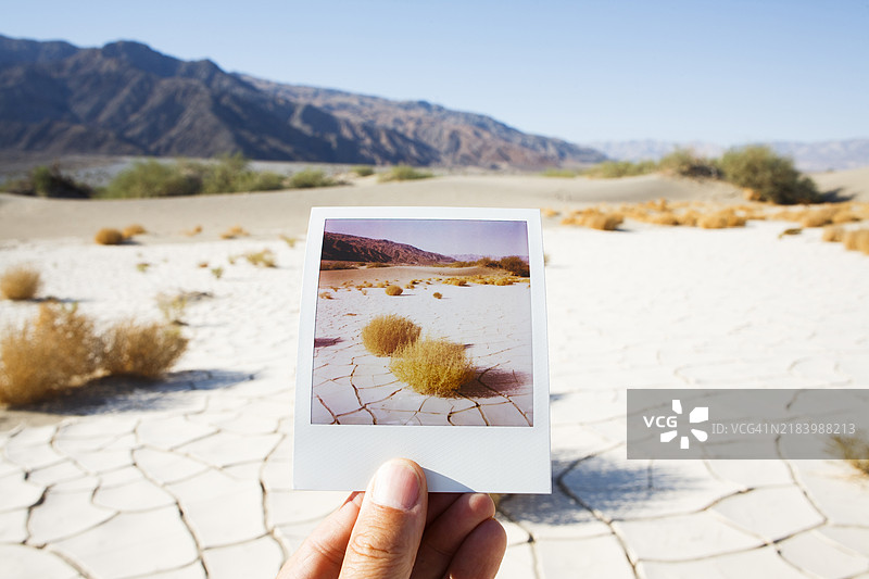 男子手持一张显示干裂泥土沙漠地面的即时胶卷照片，背景是该场景。图片素材