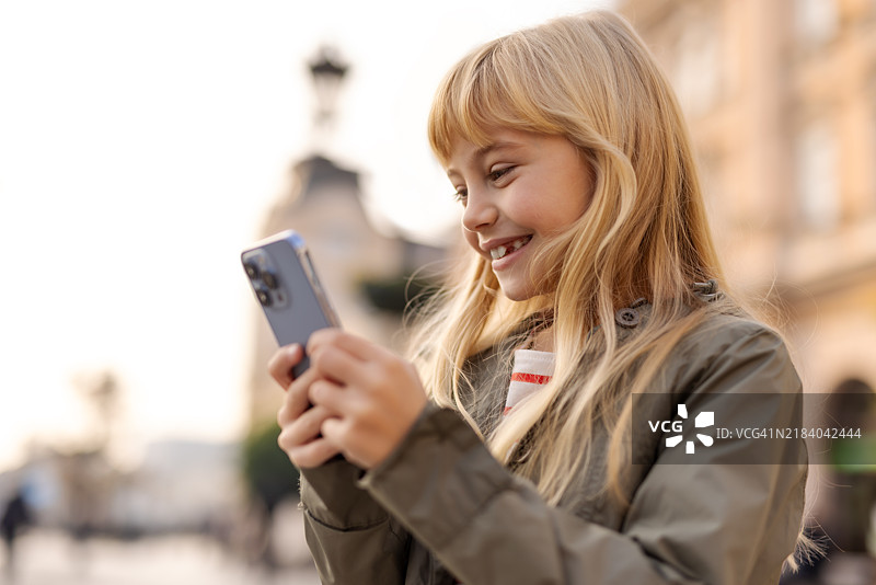 一个面带灿烂笑容的快乐小女孩在城市中心的活力氛围中与手机互动。图片素材