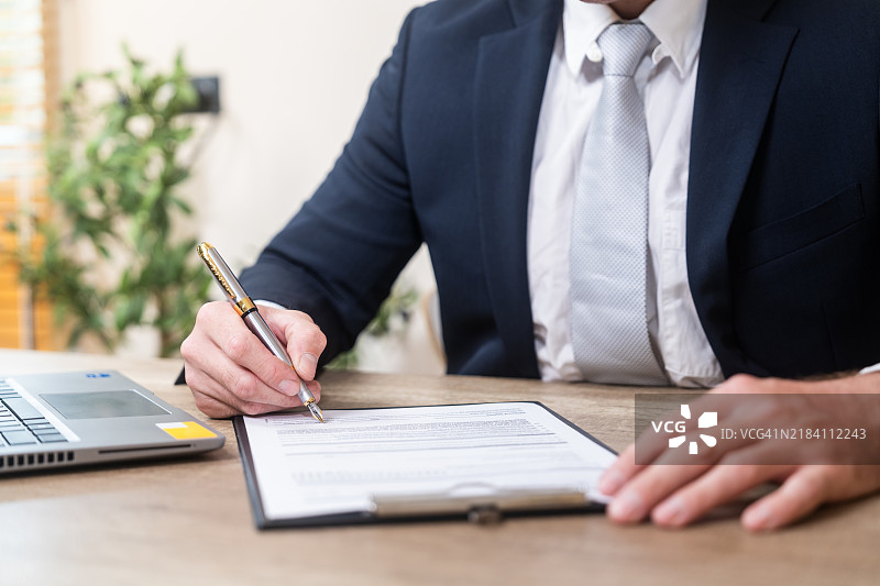商人在办公室桌子上用笔签署文件，旁边有笔记本电脑。图片素材