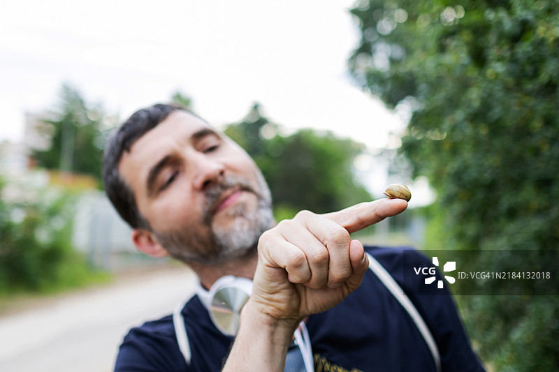 中年男子戴着耳机，享受着周围美丽自然的景色。他正在手中拿着并观察一只蜗牛。图片素材
