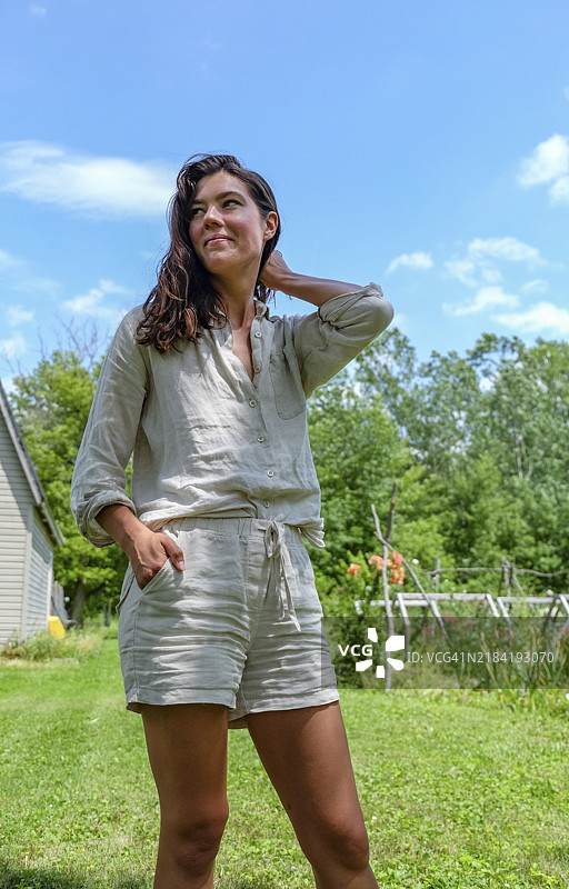 女人在阳光明媚的日子里站在花园里，一只手放在臀部，另一只手轻触脖子，微笑着仰望天空。图片素材