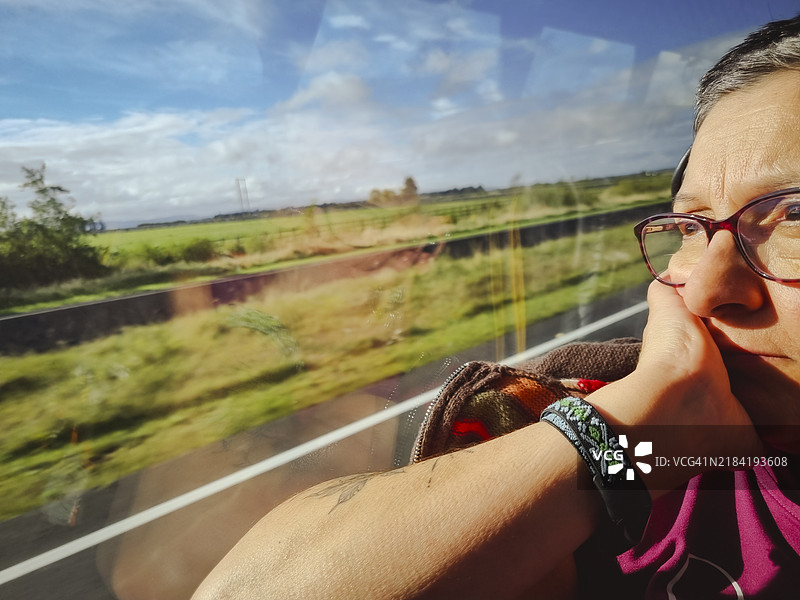 独自旅行的女性坐在窗边的公交车上，望着窗外的风景，阳光洒在她的脸上。图片素材