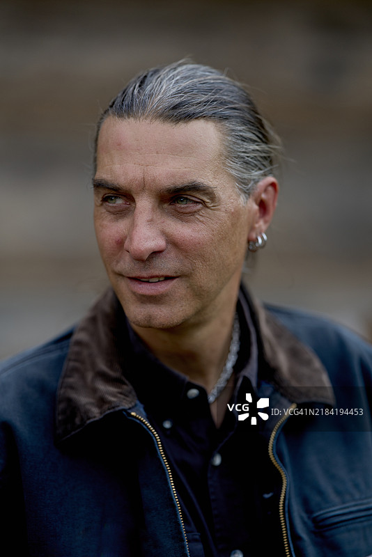 一位成熟的灰发男士穿着皮夹克，沉思地侧望，背景模糊。图片素材