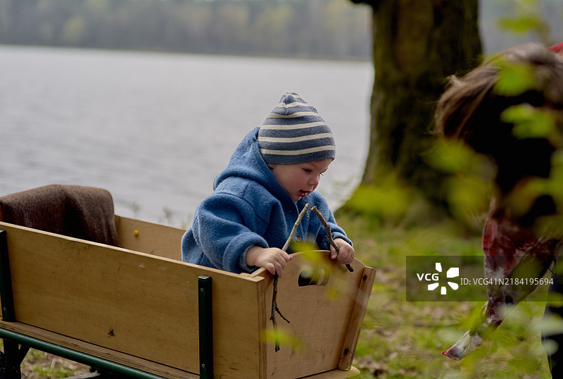 穿着蓝色外套的幼儿在湖边玩木制小车，背景是树木。图片素材
