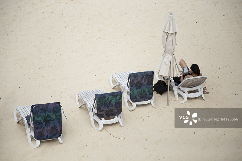 从空中俯瞰一片沙滩，一个人躺在阳光躺椅上，遮阳伞关闭，旁边是空椅子和毛巾，地点：库克群岛拉罗汤加的蒂蒂卡维卡。图片素材