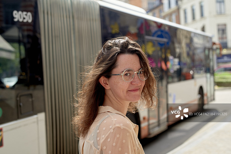 微笑的戴眼镜女性站在阳光明媚的城市街道上，面前是一辆行驶中的公交车图片素材