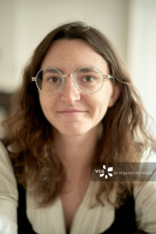 微笑的年轻女性，戴着圆形眼镜，留着波浪形的头发，穿着正式的白色衬衫，背景模糊柔和。图片素材