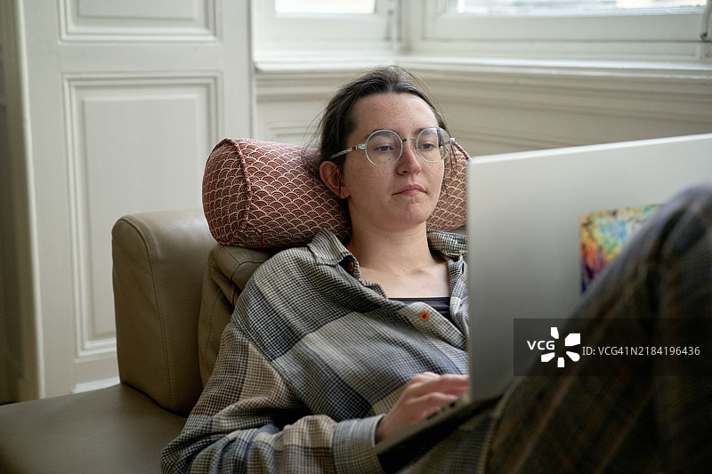 一位专注的成年人戴着眼镜，穿着格子衬衫，舒适地靠在米色扶手椅上使用笔记本电脑。图片素材