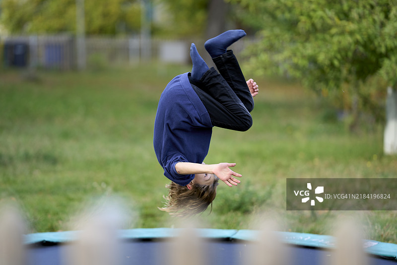 一个人在公园的绿树环绕中，在蹦床上做后空翻。图片素材