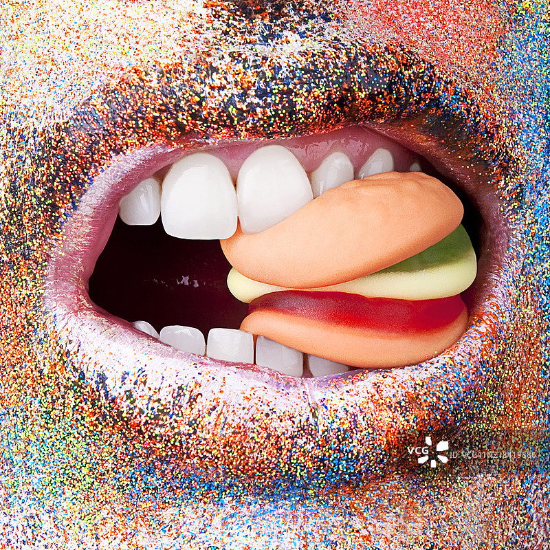 特写镜头：一张涂有五彩闪亮口红的嘴巴正在咬一堆软糖。图片素材