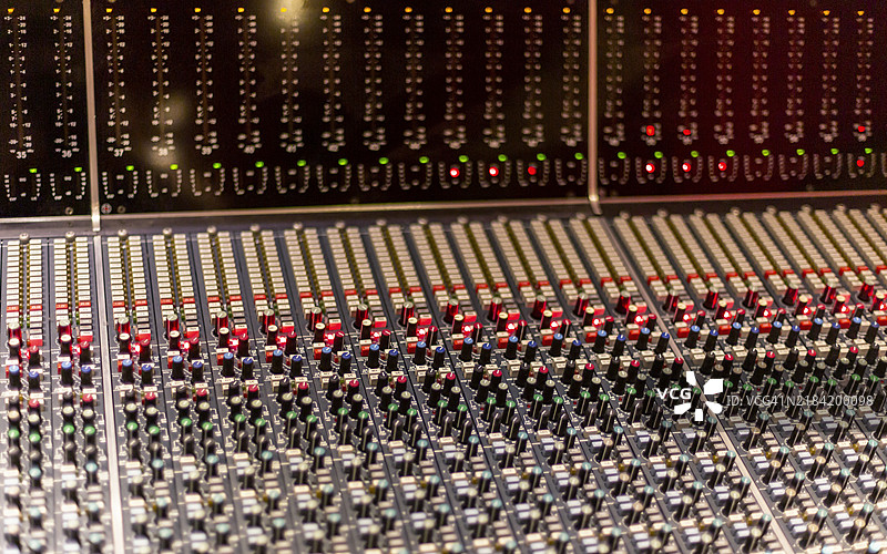 专业音频混音台的特写，配有众多调节旋钮和滑动推子，红绿指示灯闪烁。图片素材