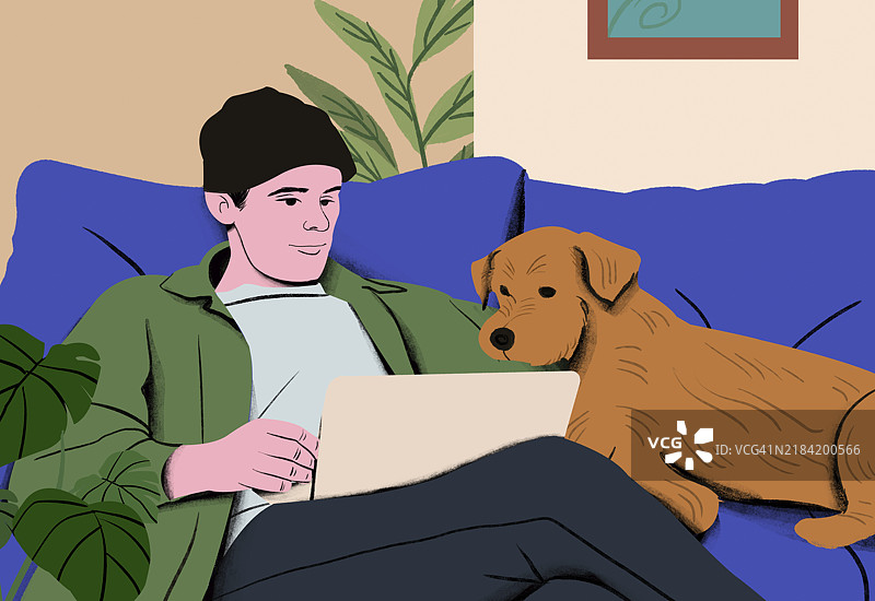 一个微笑的年轻人戴着毛线帽，穿着绿色夹克，愉快地使用着他的笔记本电脑，旁边的蓝色沙发上趴着一只棕色狗。图片素材