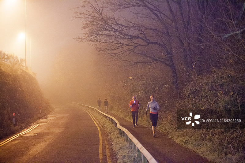 清晨的慢跑者在雾气弥漫的道路上，路旁是树木，街灯照亮了周围。图片素材