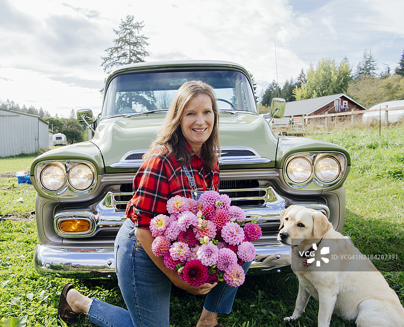 微笑的女人手捧花束，坐在乡村的复古卡车旁，身边有一只狗。图片素材