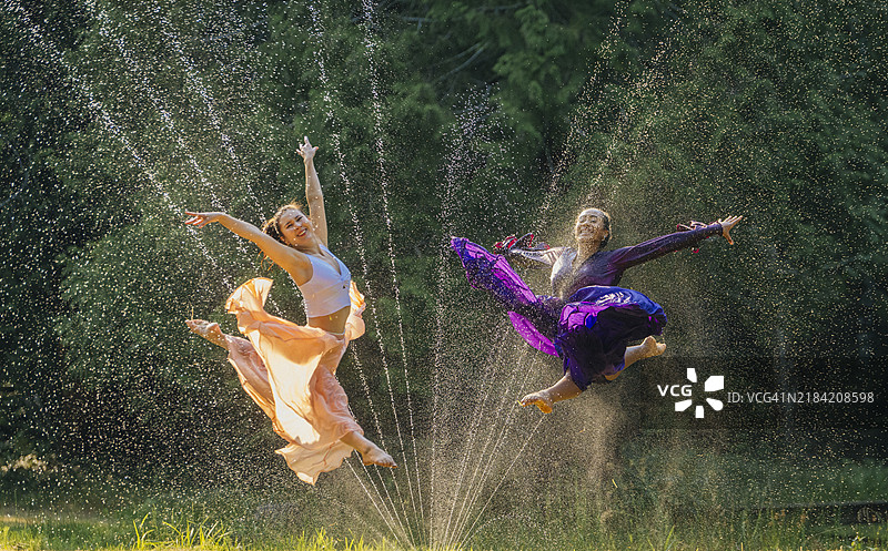 两位穿着色彩斑斓服装的舞者在水雾中跃起，背景是郁郁葱葱的绿色景色。图片素材